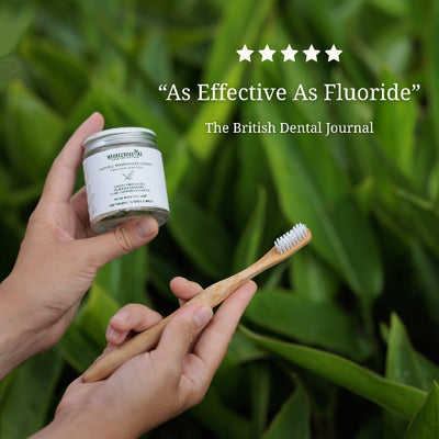 Hand holding a container of toothpaste and a bamboo toothbrush with green leaves in the background