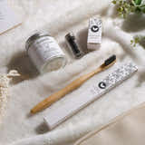 A flat-lay of a bamboo toothbrush in its white patterned packaging, a white jar candle, a small black container, and a small box, all arranged on a soft white textured blanket.