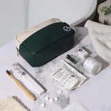 A dark green and cream zippered toiletry bag sits on a white marble surface with bath and beauty props, including a bamboo toothbrush, toothpaste tube, jar, and pouch. A folded white towel and a white vessel are visible in the background.