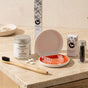 A bamboo toothbrush and several branded oral care items are arranged on a light stone countertop. Visible items include a bamboo toothbrush, a jar of natural toothpaste, a box of activated charcoal, a small box of charcoal packets, a clear tray, an orange-red mouthpiece, and a clear retainer or mouthguard resting in the tray.