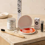 A bamboo toothbrush and several branded oral care items are arranged on a light stone countertop. Visible items include a bamboo toothbrush, a jar of natural toothpaste, a box of activated charcoal, a small box of charcoal packets, a clear tray, an orange-red mouthpiece, and a clear retainer or mouthguard resting in the tray.
