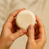 Hand holding a round, coconut shampoo against a beige background